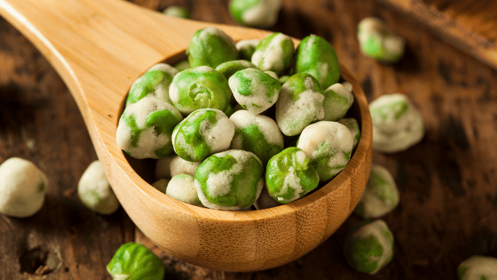 A wooden spoon filled with wasabi-coated peas rests on a rustic wooden surface. Several wasabi peas are scattered around the spoon. The peas are green with a white, powdery wasabi coating.