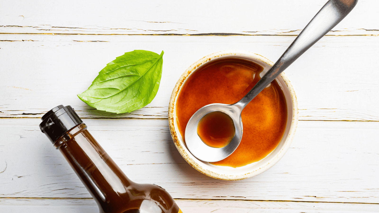 A bowl of brown sauce with a spoon in it sits on a white wooden surface. Next to the bowl is a fresh green basil leaf. A dark glass bottle is partially visible in the bottom left corner of the image.