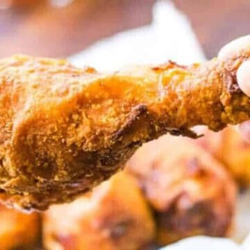 A close-up of a hand holding a crispy fried chicken drumstick, with other pieces of chicken visible in the background on a plate.