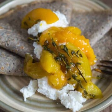 A plate with folded buckwheat crepes is topped with a dollop of ricotta cheese, peach slices, and fresh thyme. A fork lies on the right side of the plate.