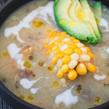 A bowl of ajiaco colombiano soup with chicken, corn on the cob, and avocado.