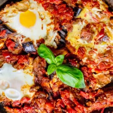 A skillet containing a dish of shakshuka is shown. The dish features poached eggs in a sauce of tomatoes, chili peppers, and onions, garnished with fresh basil leaves. The vibrant colors of the ingredients create a visually appealing presentation.