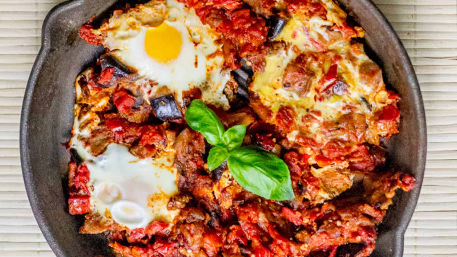 A skillet containing a dish of shakshuka is shown. The dish features poached eggs in a sauce of tomatoes, chili peppers, and onions, garnished with fresh basil leaves. The vibrant colors of the ingredients create a visually appealing presentation.