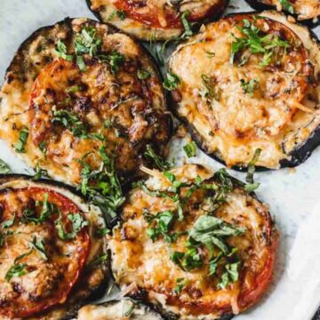 A plate of cooked eggplant slices topped with melted cheese, tomato, and chopped green herbs is shown. This quick and easy dinner rests on a white oval plate placed on a wooden surface with a dark cloth napkin nearby.