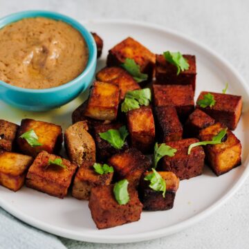 A white plate with cubed tofu garnished with cilantro, served alongside a small bowl of sauce, perfect for Cinco De Mayo recipes. The tofu is browned and arranged neatly on the plate. There's a light blue cloth napkin to the left of the plate.