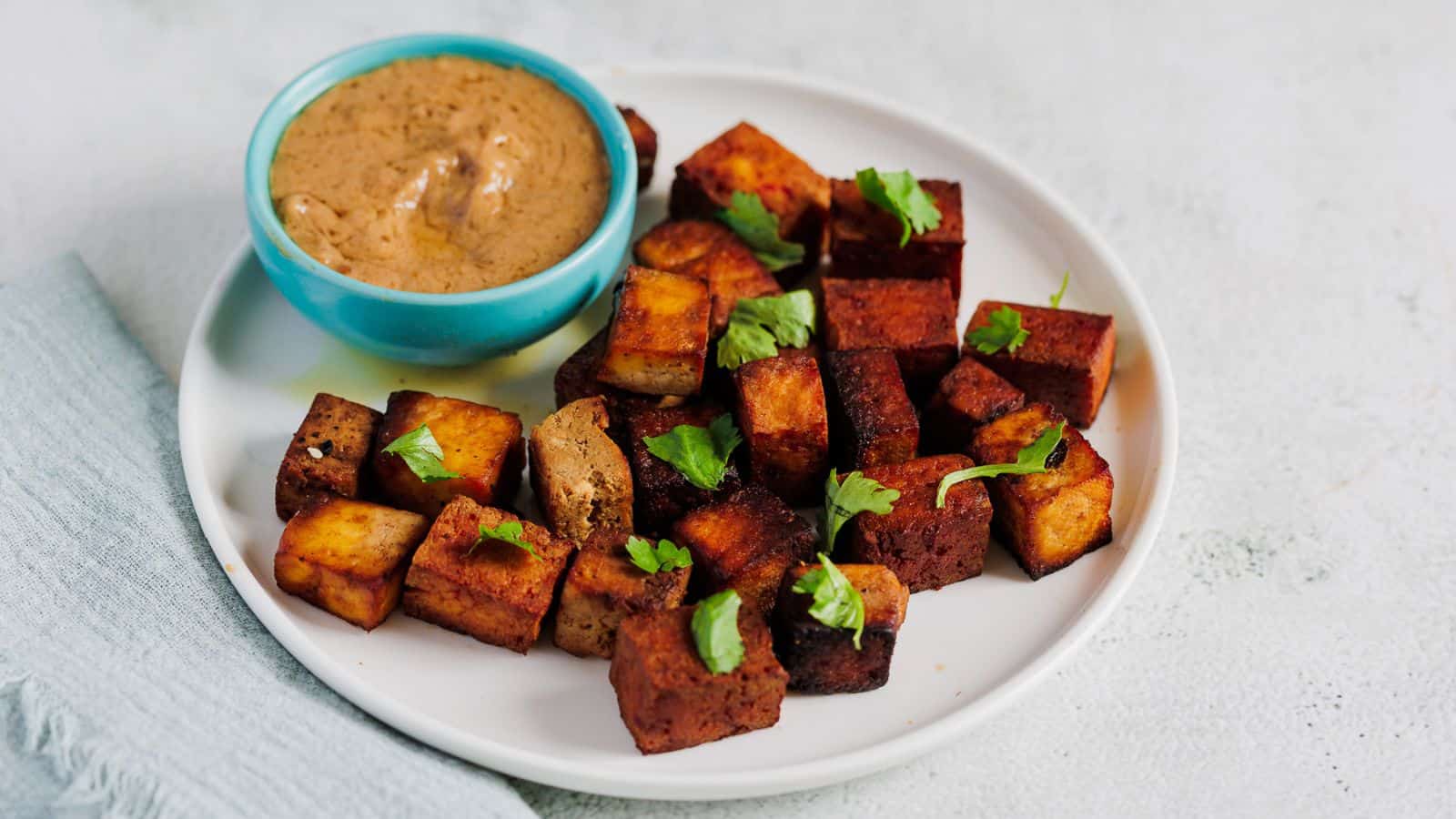 A white plate with cubed tofu garnished with cilantro, served alongside a small bowl of sauce, perfect for Cinco De Mayo recipes. The tofu is browned and arranged neatly on the plate. There's a light blue cloth napkin to the left of the plate.