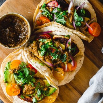 A wooden platter holds three open tacos filled with spiced meat, sliced tomatoes, cucumbers, onions, and topped with fresh cilantro, served with a side of dark sauce in celebration of