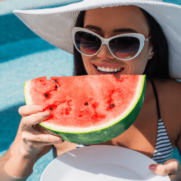 A woman wearing a sun hat, sunglasses, and a striped swimsuit is standing in a pool while holding and eating a slice of watermelon. She is smiling and appears to be enjoying the sunny weather.