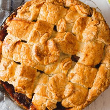 A golden-brown, lattice-crust pie sits on a white wooden surface. The pie filling appears to be apple-based, with a slightly visible edge of the filling around the crust. Beside the pie is a gray cloth and a bowl containing green apples and apple slices.