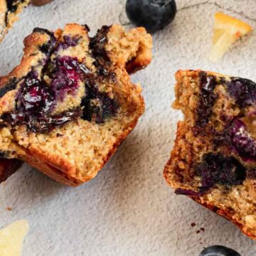 Close-up of blueberry breakfast breads on a light surface, some broken open showing juicy berries inside. Scattered blueberries and crumbs are visible around them.
