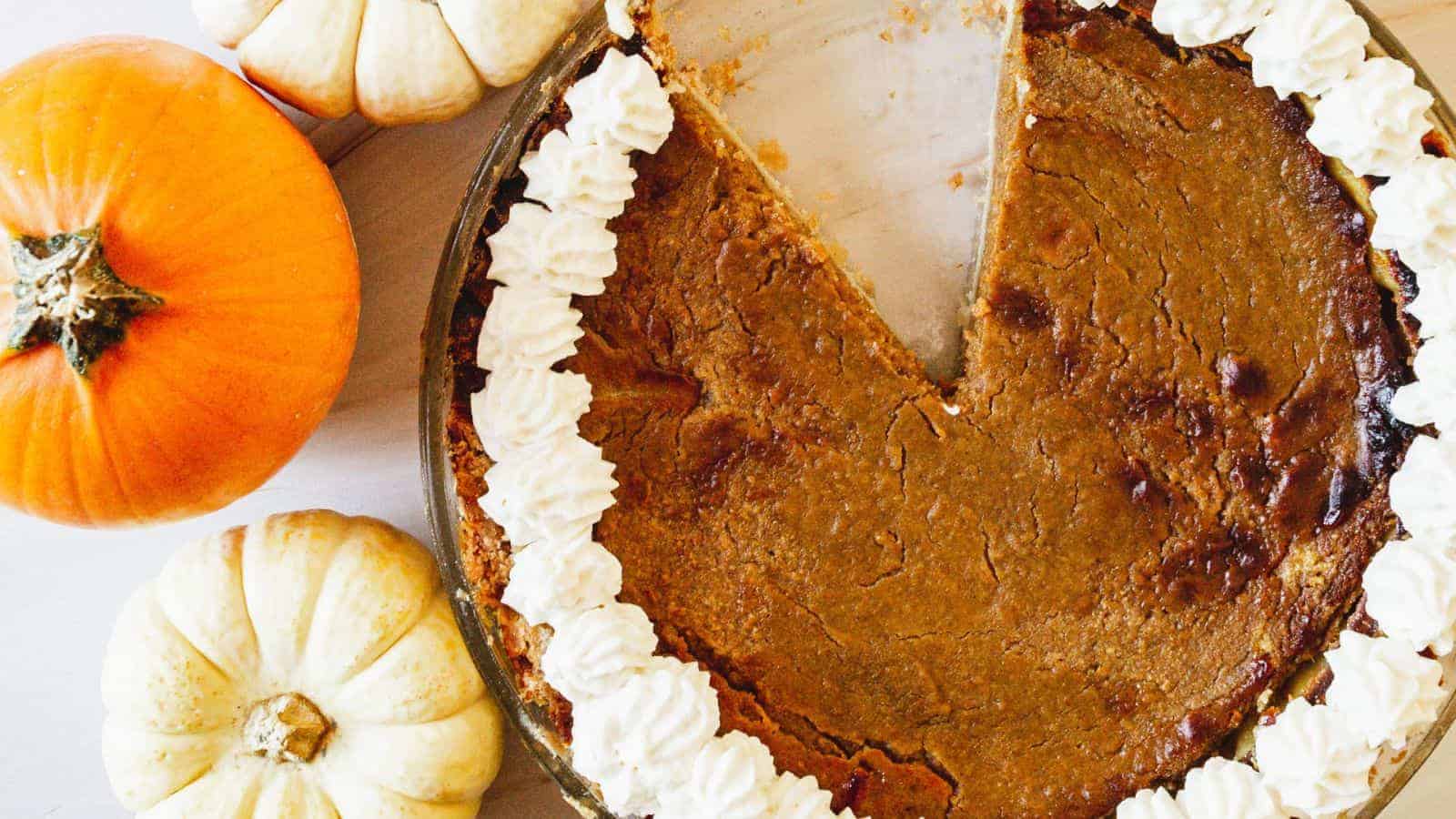 A pumpkin pie with a slice missing is surrounded by dollops of whipped cream along the edge. The pie is placed on a light-colored surface, and there are three small pumpkins next to it: one orange and two white. Perfect for dessert lovers who savor retro recipes!