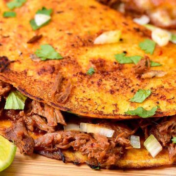 Close-up of a crispy taco filled with shredded beef, garnished with chopped onions and cilantro, served with lime wedges on a wooden surface. Ideal for Cinco de Mayo recipes.
