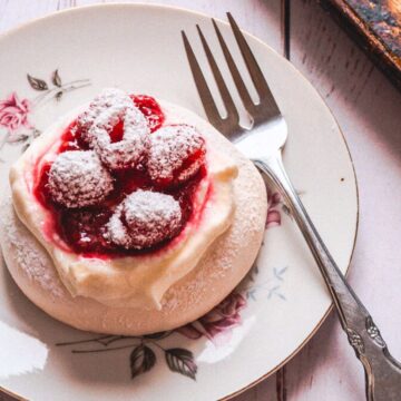 A dessert plate with a floral design holds a small pavlova topped with whipped cream, raspberries, and powdered sugar—a true dessert classic making a delightful comeback. A silver fork rests on the right side of the plate, which sits on a light wooden surface with a cloth napkin to the left and a cutting board to the right.