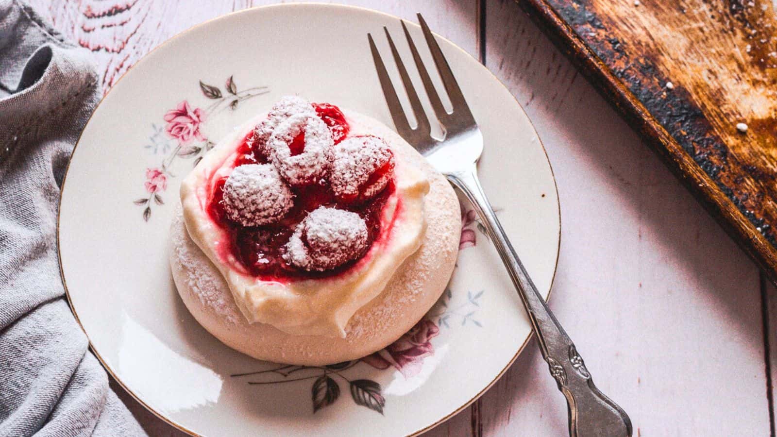 A dessert plate with a floral design holds a small pavlova topped with whipped cream, raspberries, and powdered sugar&mdash;a true dessert classic making a delightful comeback. A silver fork rests on the right side of the plate, which sits on a light wooden surface with a cloth napkin to the left and a cutting board to the right.