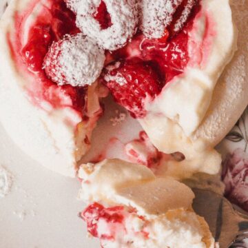 A plate with a dessert classic consisting of a meringue-like base topped with whipped cream, red strawberries, and powdered sugar. A fork lies next to the dessert, with a piece taken off. The plate has a floral design.
