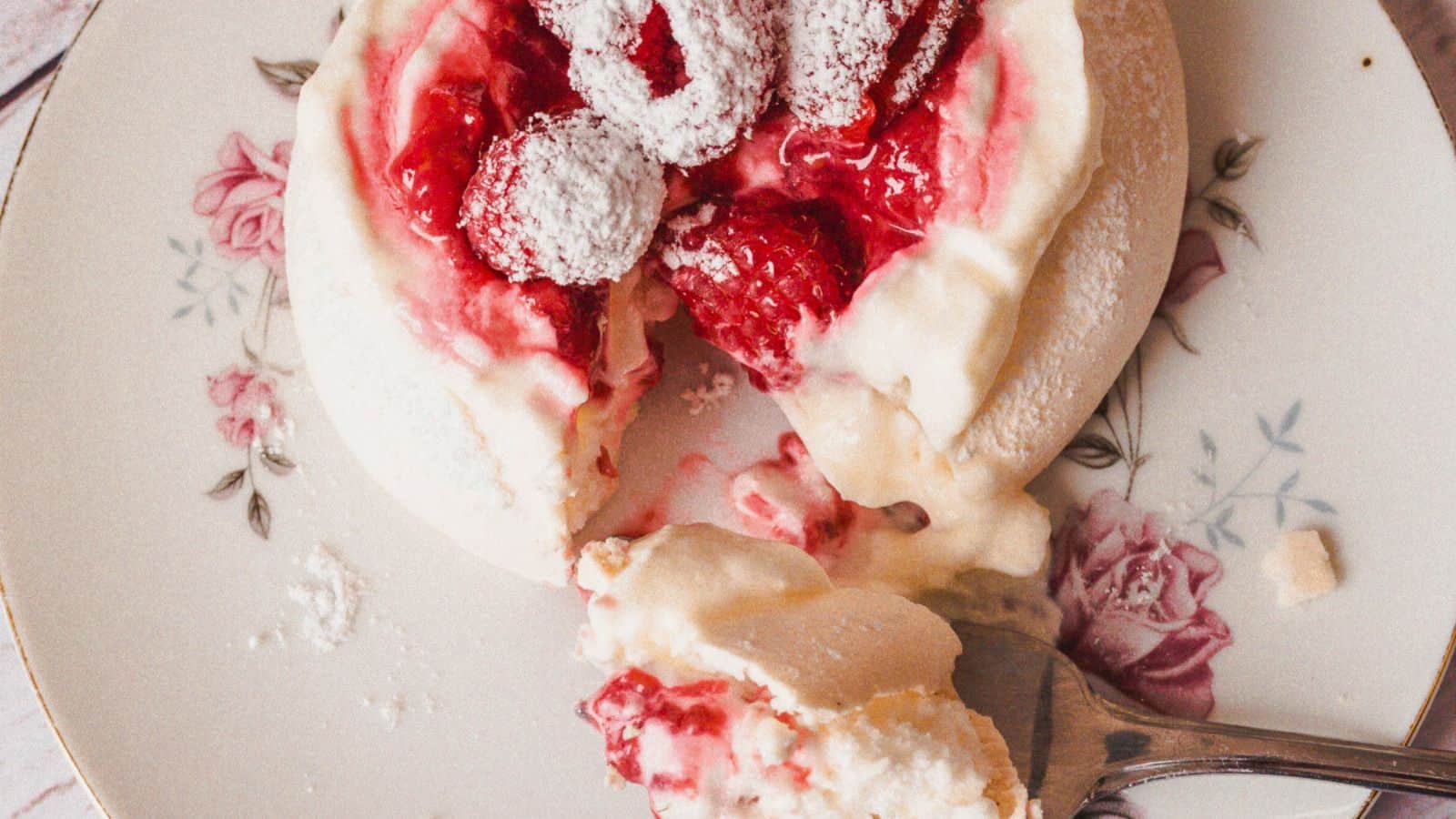 A plate with a dessert classic consisting of a meringue-like base topped with whipped cream, red strawberries, and powdered sugar. A fork lies next to the dessert, with a piece taken off. The plate has a floral design.