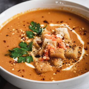 A bowl of creamy bell pepper bisque garnished with croutons, chopped parsley, diced tomatoes, and a drizzle of cream, served on a dark surface.