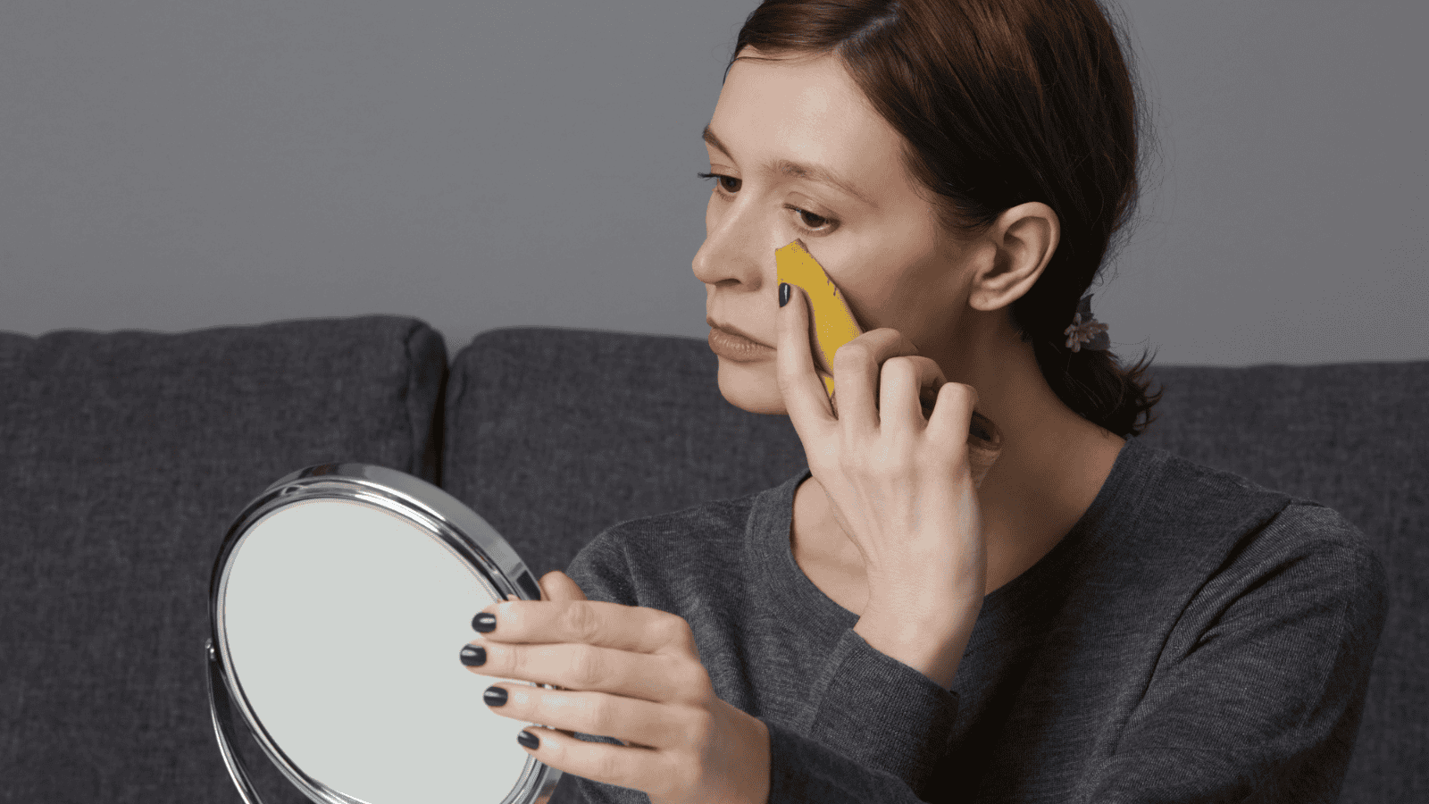 A person with a neutral expression uses a banana peel on their face while looking into a handheld mirror. The person has dark hair and is wearing a gray shirt. The background is a dark gray couch against a light gray wall.