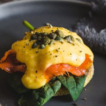 A close-up image of a retro Mother's Day eggs benedict with smoked salmon and spinach, topped with hollandaise sauce and capers, served on a dark round plate with a fork on the side.