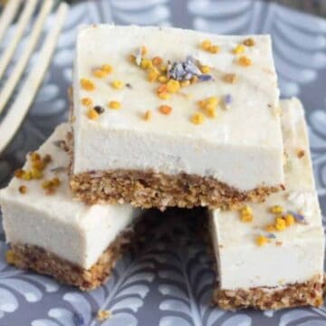 Three pieces of layered dessert bars are stacked on a decorative grey and white plate. The bars have a crumbly base with a creamy topping, garnished with small yellow and purple sprinkles. A fork is placed beside the bars on the plate, inviting you to dig in without hesitation or an auto draft of appetite!