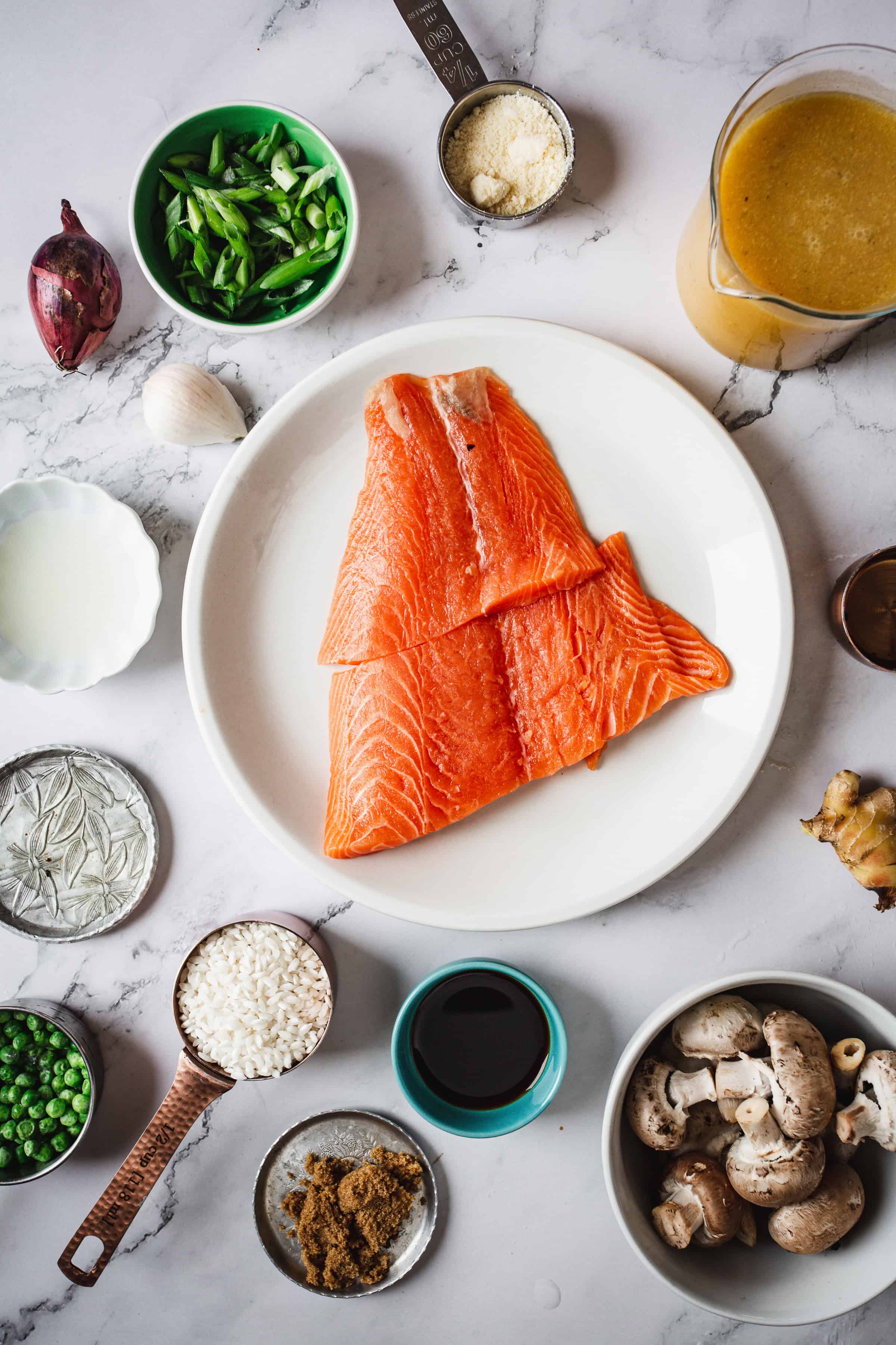 A top view of a cooking setup with ingredients arranged around a white plate of raw salmon fillets, perfect for a Teriyaki Glazed Salmon recipe. Ingredients include peas, sliced green onions, rice for risotto, mushrooms, soy sauce, ginger, a small onion, garlic, and a cup of broth. All items are placed on a marble surface.