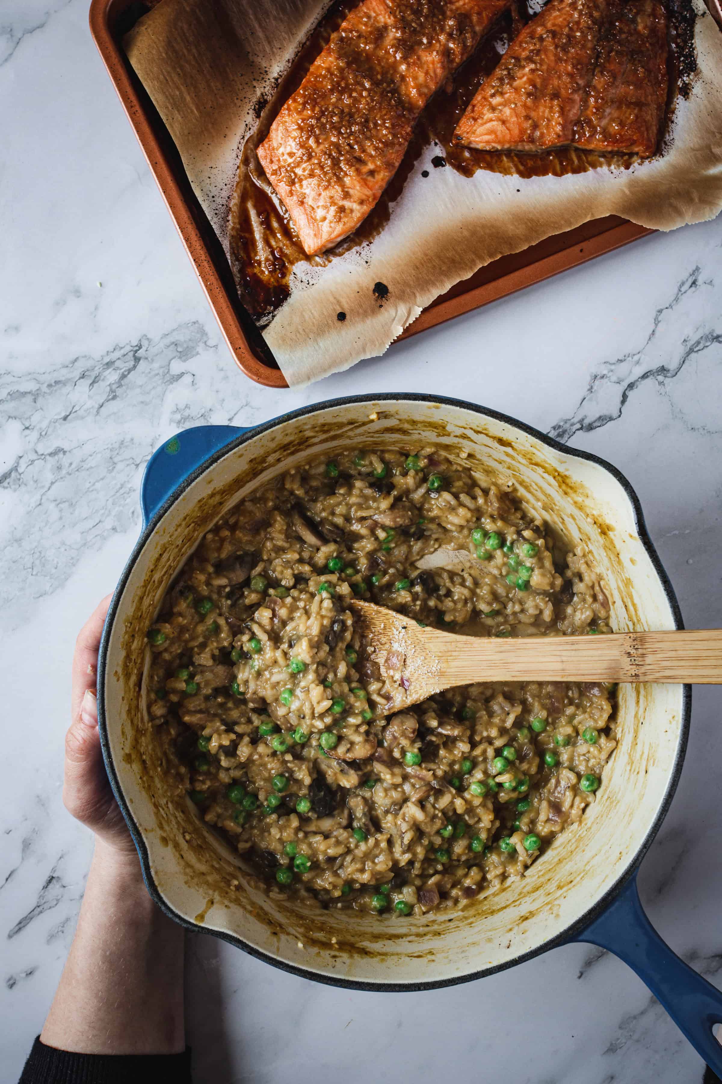 A hand holds a blue pot containing risotto with peas and mushrooms, while a wooden spoon rests in the pot. To the side, a baking pan with parchment paper showcases two pieces of Teriyaki Glazed Salmon. The scene is set on a marble countertop.