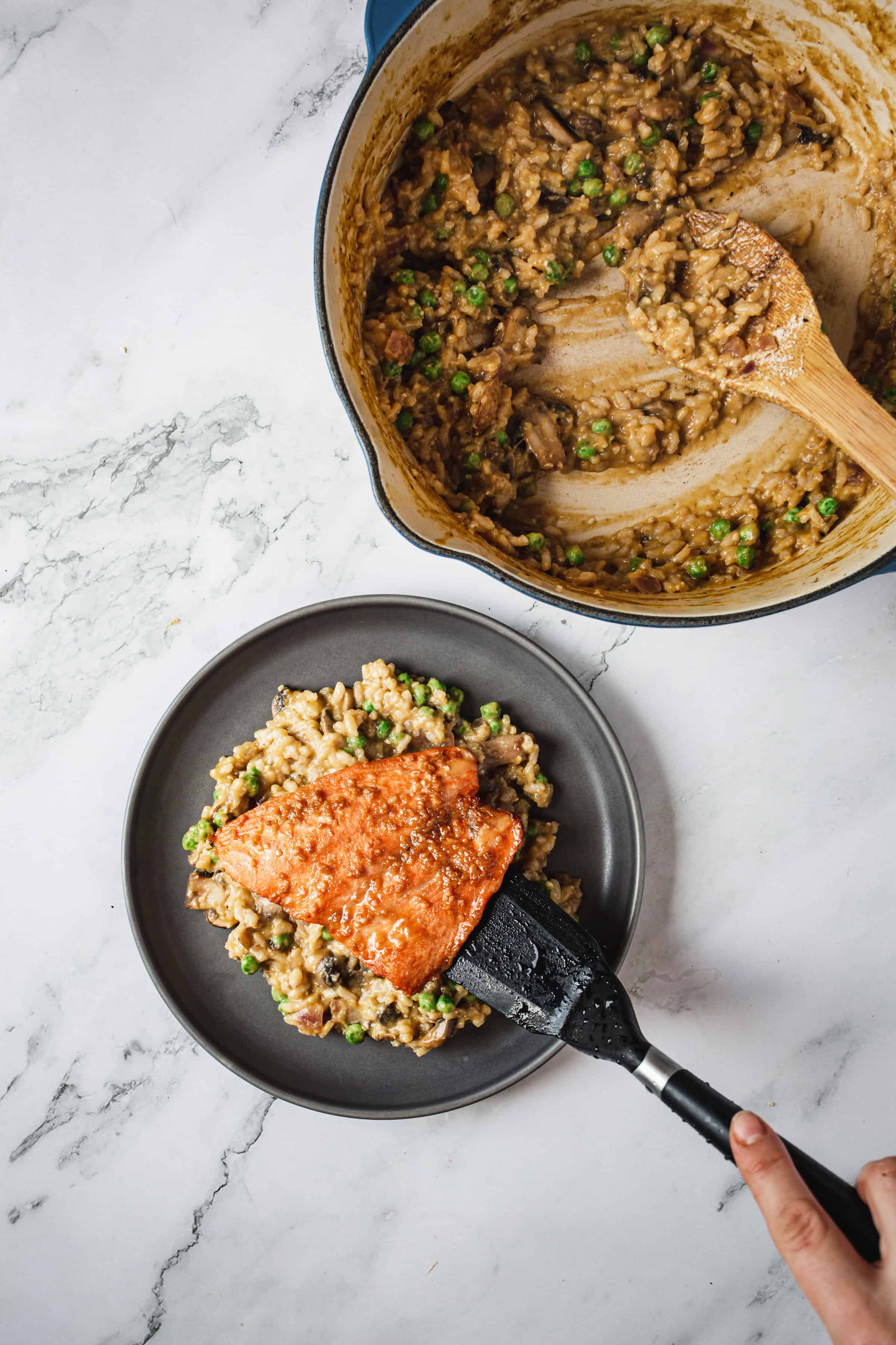 Top-down view of a person placing a piece of teriyaki glazed salmon onto a plate of risotto with peas using a black spatula. The pot with remaining risotto is visible in the background with a wooden spoon inside. The scene is set on a white marble surface.