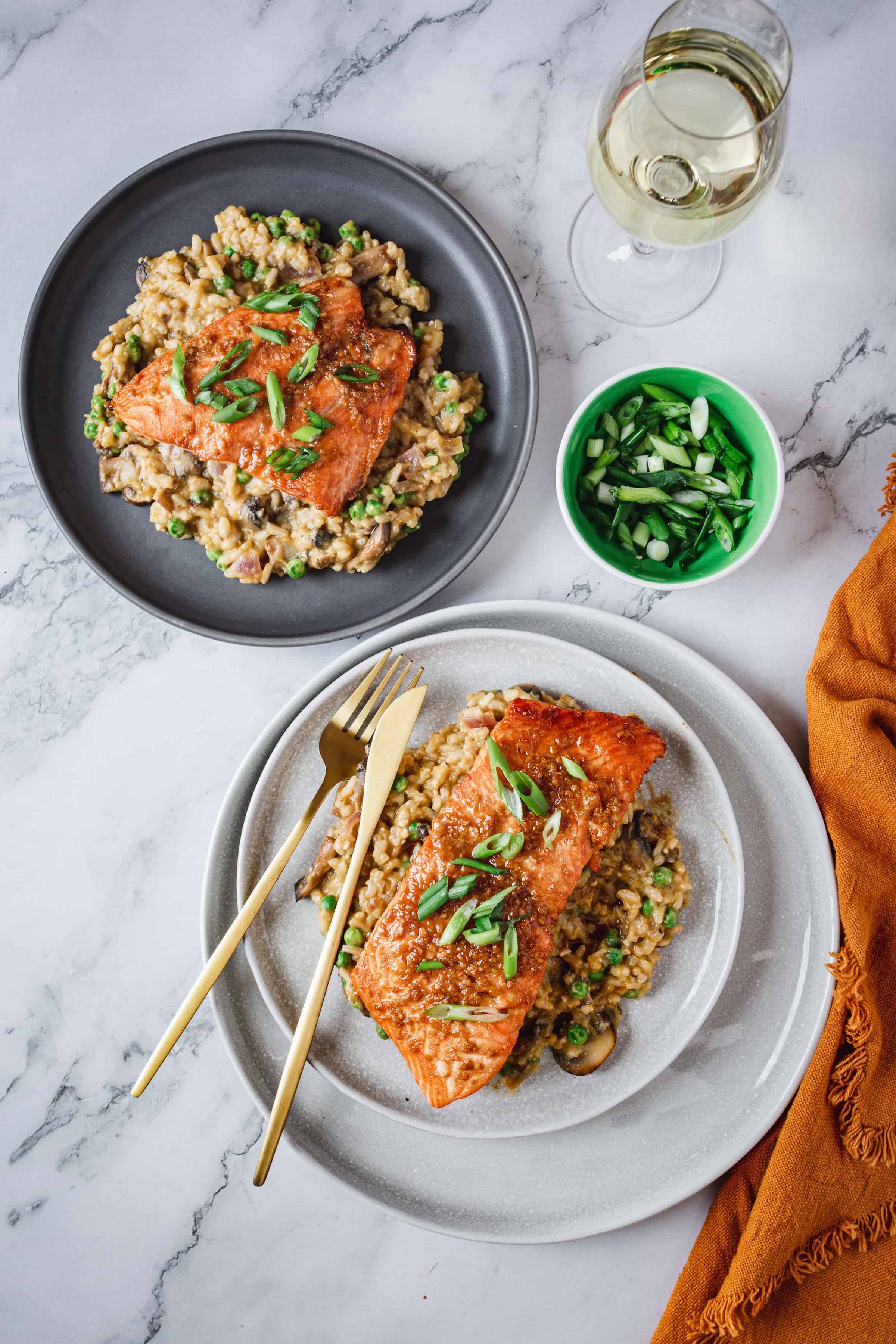 Two plates of Teriyaki Glazed Salmon over mushroom and pea risotto garnished with green onions. Golden cutlery is on the plate at the bottom. A small bowl of green onions and a glass of white wine are next to the plates on a marble surface with an orange cloth—an enticing risotto recipe for any occasion.