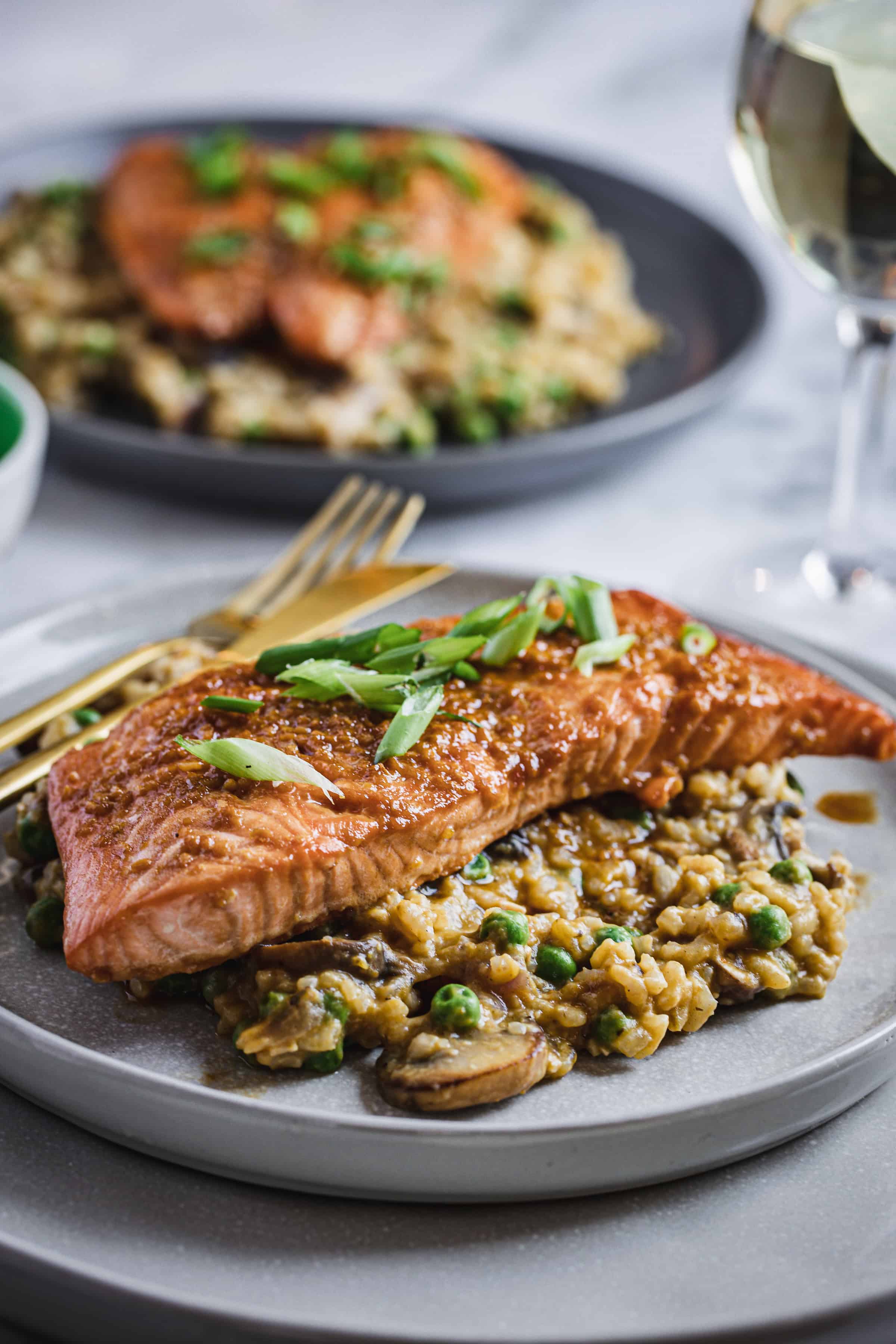 A plated meal features a seared teriyaki glazed salmon fillet topped with green onions, resting on a bed of mushroom risotto with peas. Behind, a similar dish is visible out of focus. A fork and knife are placed beside the plate, and a glass of white wine is in the background.