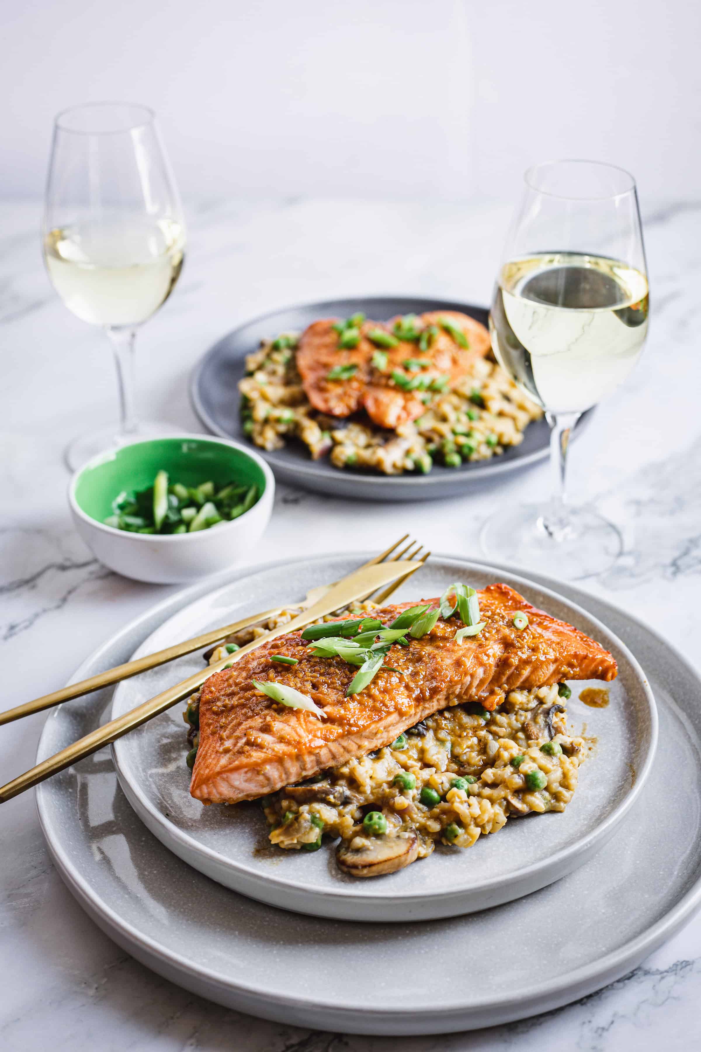 A dinner setting features two plates of risotto topped with Teriyaki Glazed Salmon and sliced green onions. Two glasses of white wine sit beside the plates. A small green bowl containing additional green onions rests in the background on a white marble surface.