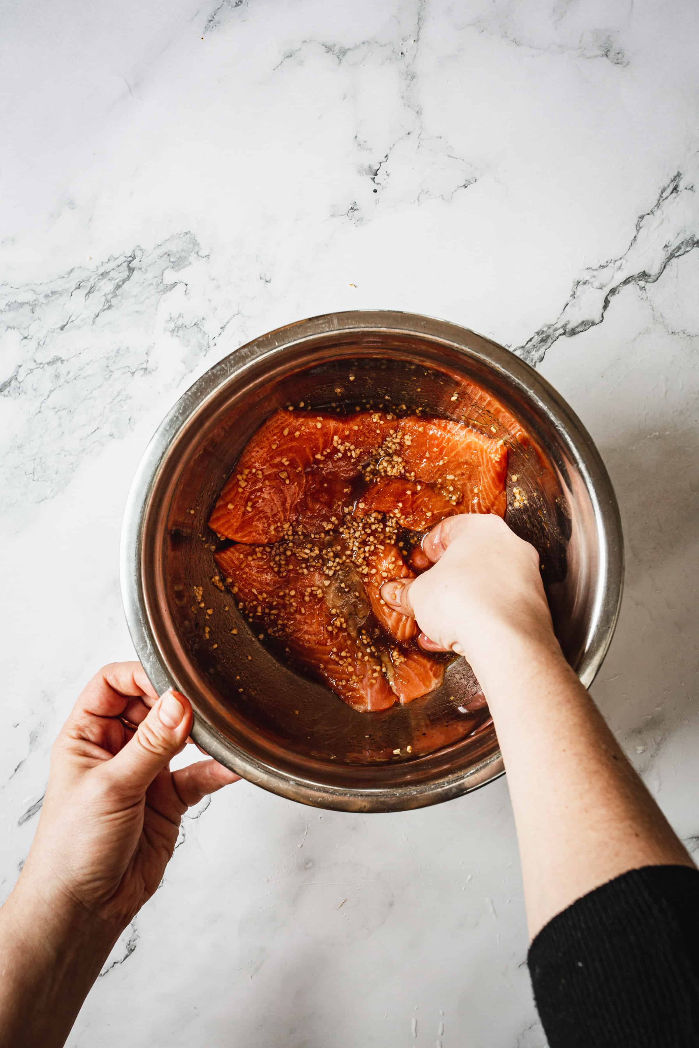 A person is preparing marinated teriyaki glazed salmon in a stainless steel bowl. The salmon appears to be seasoned with herbs and spices. The person's hands are visible, with one hand holding the bowl and the other arranging the salmon. The background is a marble surface.