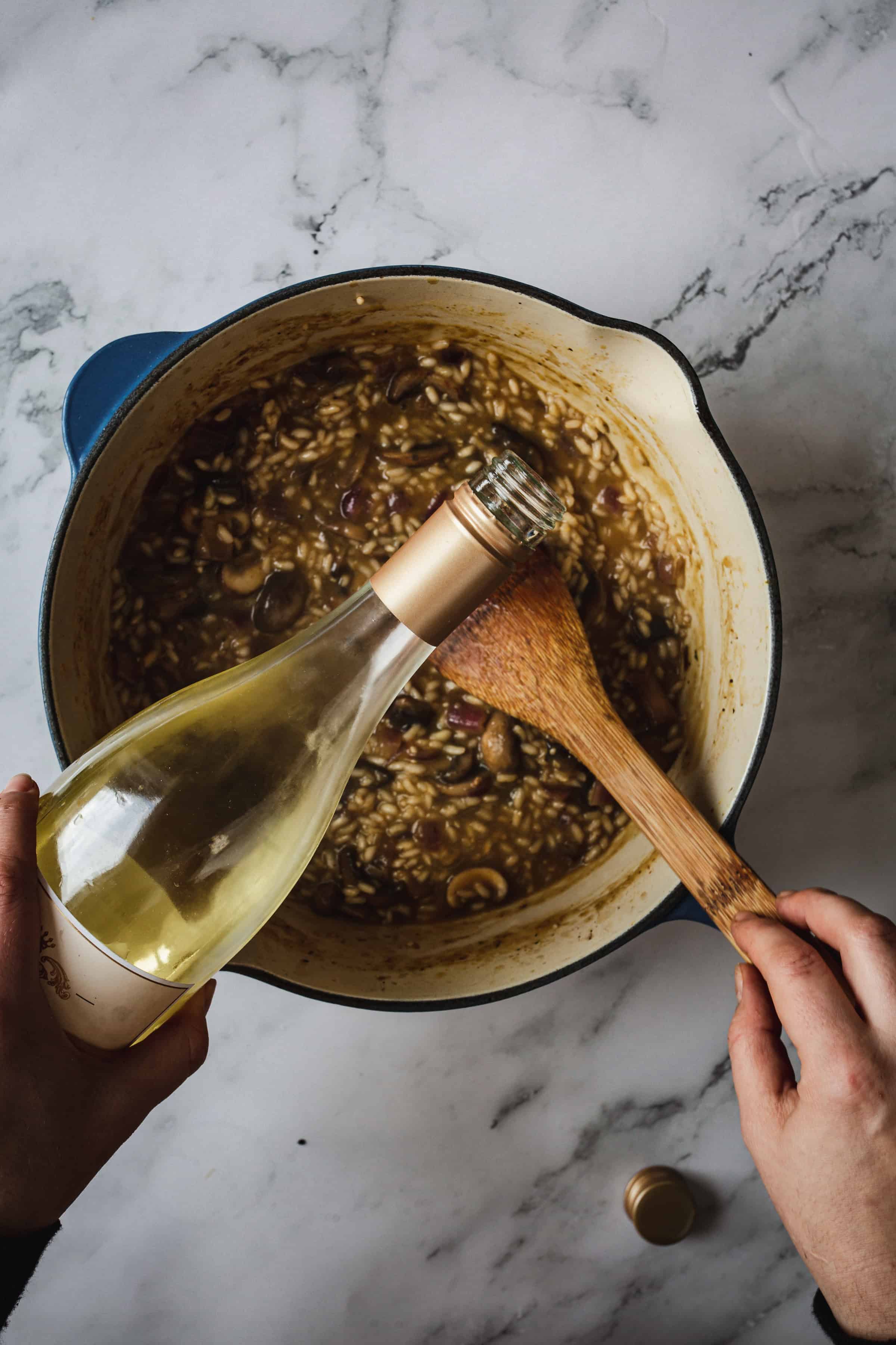 A person is holding a bottle of white wine and pouring some into a pot filled with a mushroom risotto mixture. They are also stirring the risotto with a wooden spoon. The pot, perfect for any risotto recipe, is placed on a white marble countertop.