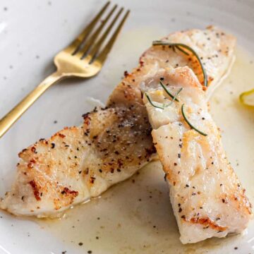 A plate features two fillets of grilled white fish, seasoned with pepper and garnished with herbs. A gold fork is placed to the upper left of the fish. Two lemon wedges are on the plate's right side. The dish is served on a white plate with a light sauce.