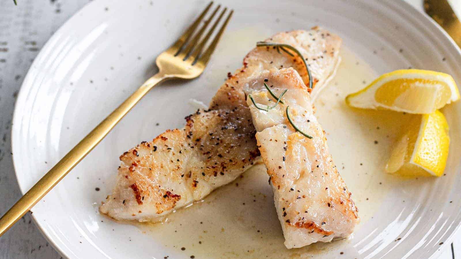 A plate features two fillets of grilled white fish, seasoned with pepper and garnished with herbs. A gold fork is placed to the upper left of the fish. Two lemon wedges are on the plate's right side. The dish is served on a white plate with a light sauce.