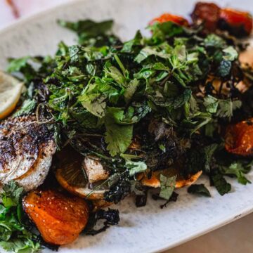 A grilled whole fish garnished with fresh herbs and surrounded by roasted cherry tomatoes is served on a white oval plate.