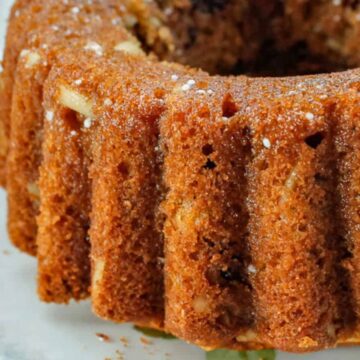 The image shows a close-up of a bundt cake on a white plate. The cake appears to be baked to a golden brown color with a textured exterior. It has visible pieces of nuts and possibly dried fruits within. The white plate has a decorative edge.