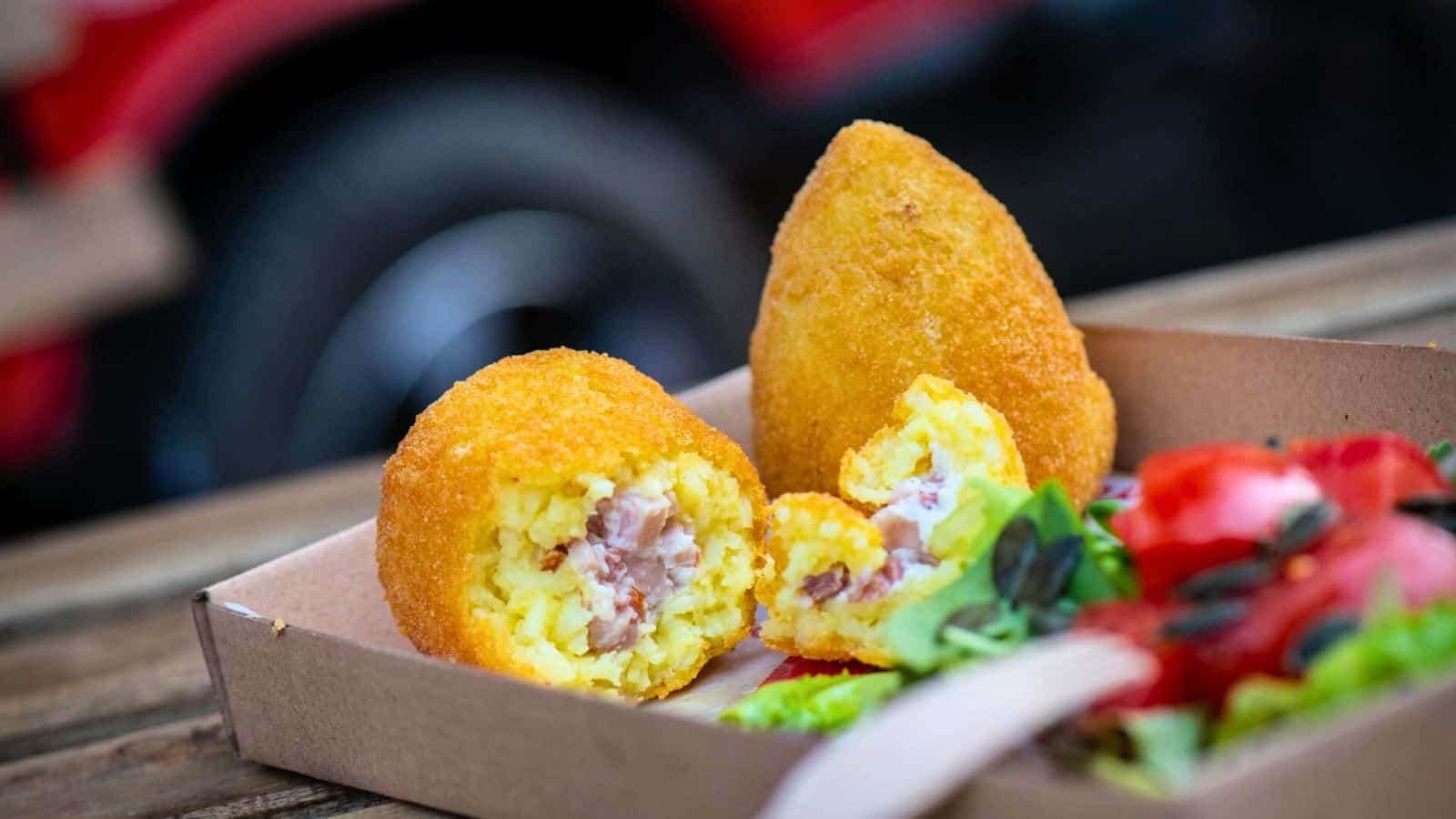 A close-up of two fried, breaded street foods, with one split open to reveal a filling of rice and bits of meat. Next to them is a side salad with a mix of greens and slices of tomato, served in a cardboard tray. The background is blurry with part of an iconic vehicle visible.