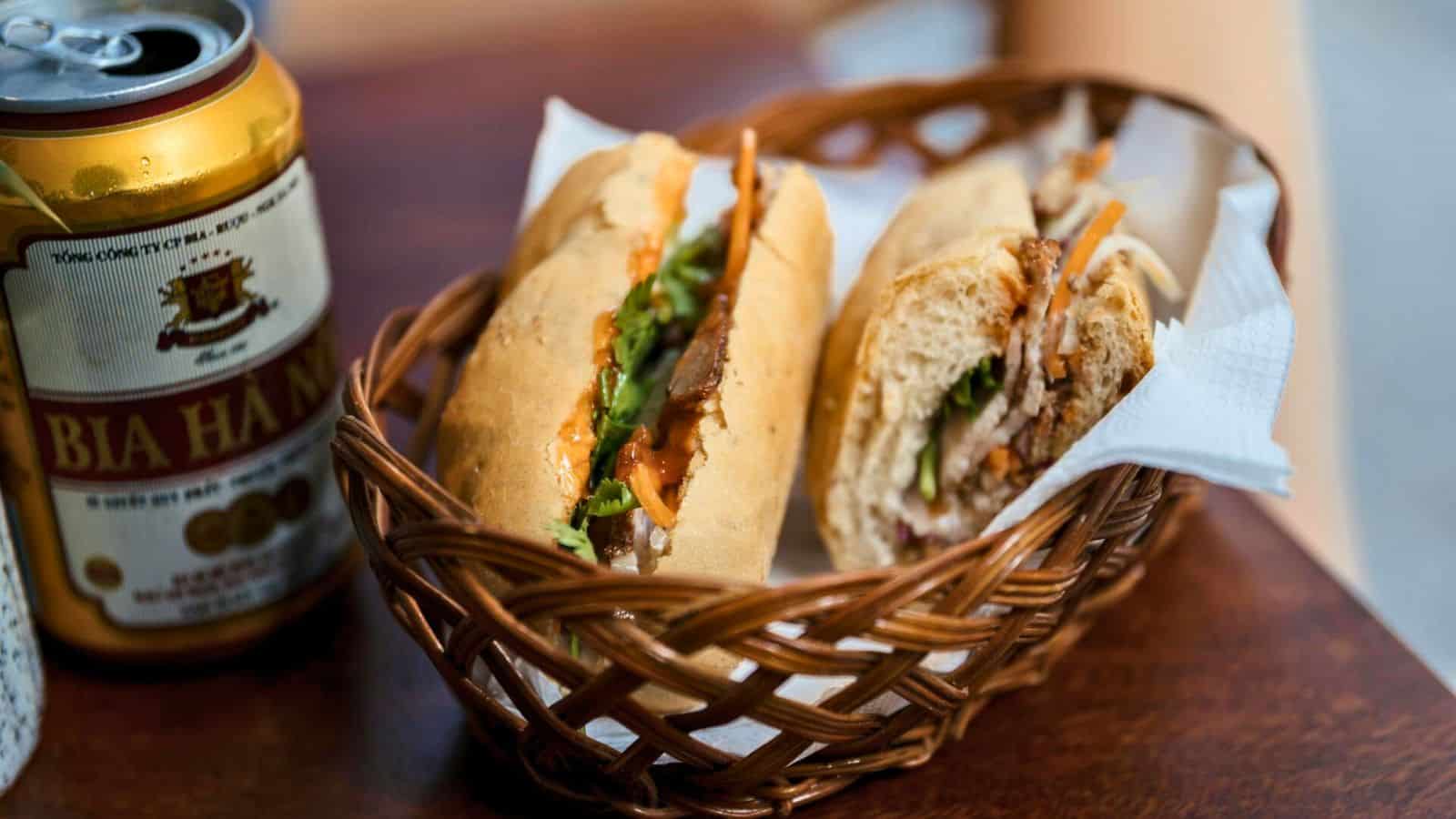 A wicker basket holding two halves of a sandwich filled with various ingredients sits on a wooden table. Beside the basket is a can of Bia Ha Noi beer.