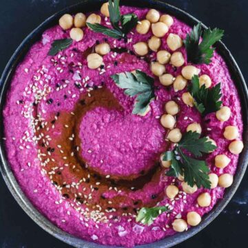 A bowl of vibrant pink beet hummus garnished with chickpeas, parsley leaves, black and white sesame seeds, and a drizzle of olive oil, placed on a dark surface.