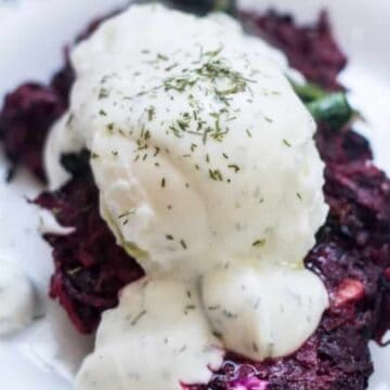 A white plate with a serving of beetroot fritters topped with a poached egg and white sauce, garnished with dill. A fork is placed to the right of the plate, and the edge of the plate has a decorative blue border.