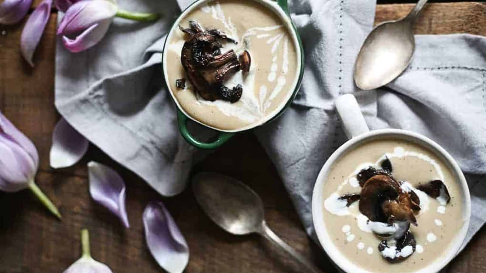 Two cups of creamy mushroom soup, a beloved offering in international dishes, sit on a wooden table. Each is topped with sautéed mushrooms and a swirl of cream. Silver spoons rest beside them, scattered purple flower petals, and a gray cloth napkin complete the setting.