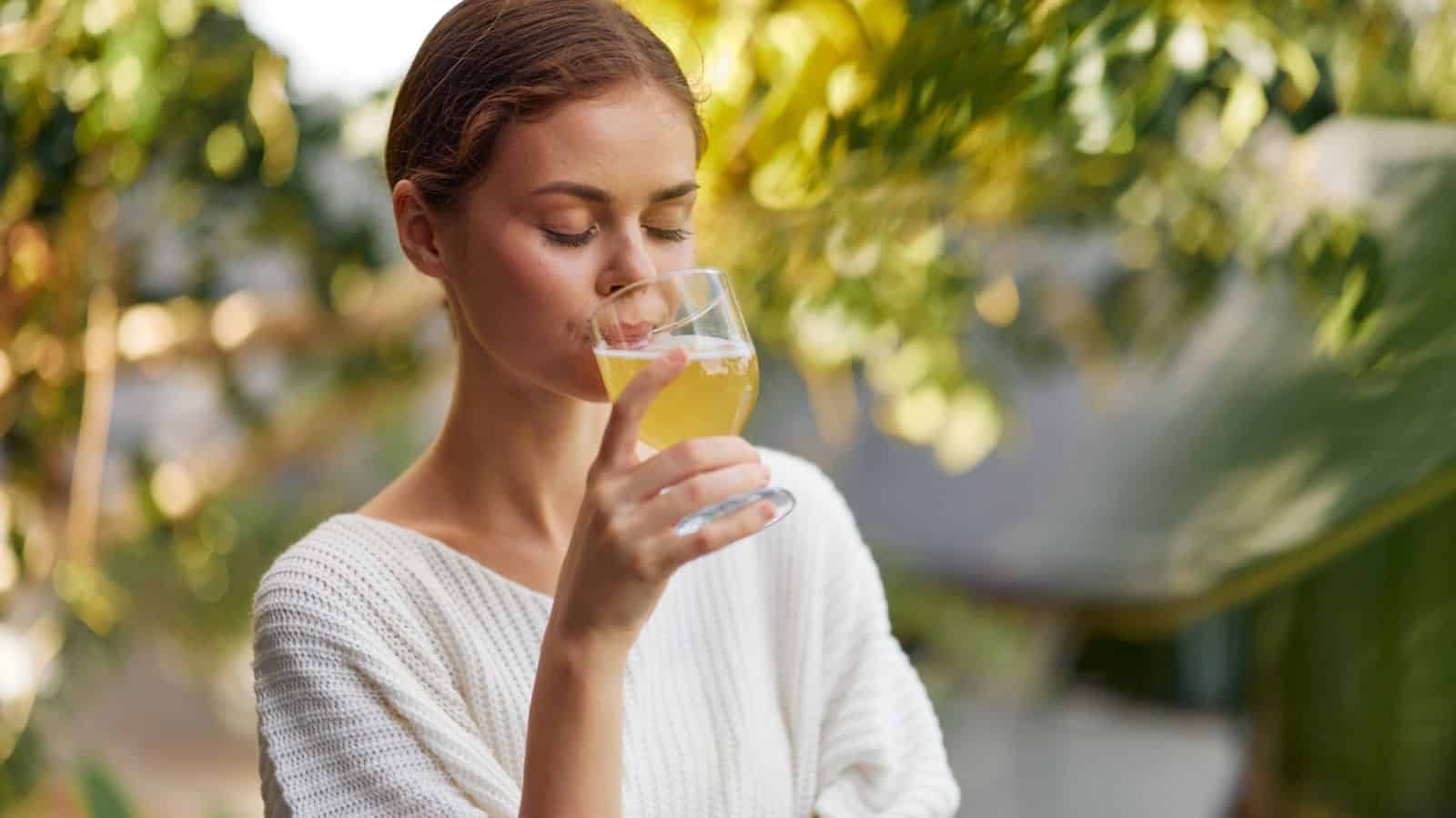A person with brown hair tied back is standing outdoors, savoring a yellow drink in a clear glass, eyes closed. The background is blurred with green foliage and sunlight filtering through.