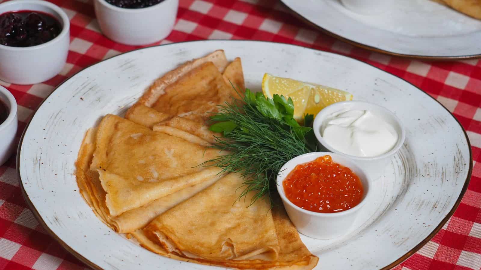 A plate of crepes garnished with sprigs of dill, a lemon wedge, a small bowl of sour cream, and red caviar sits on a red and white checkered tablecloth.