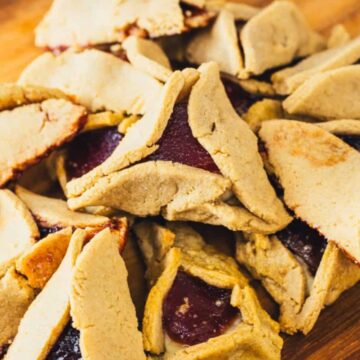 A pile of triangular-shaped cookies filled with a reddish fruit filling is displayed on a wooden surface. The cookies have a golden, slightly browned crust, with some filling visible in the openings.