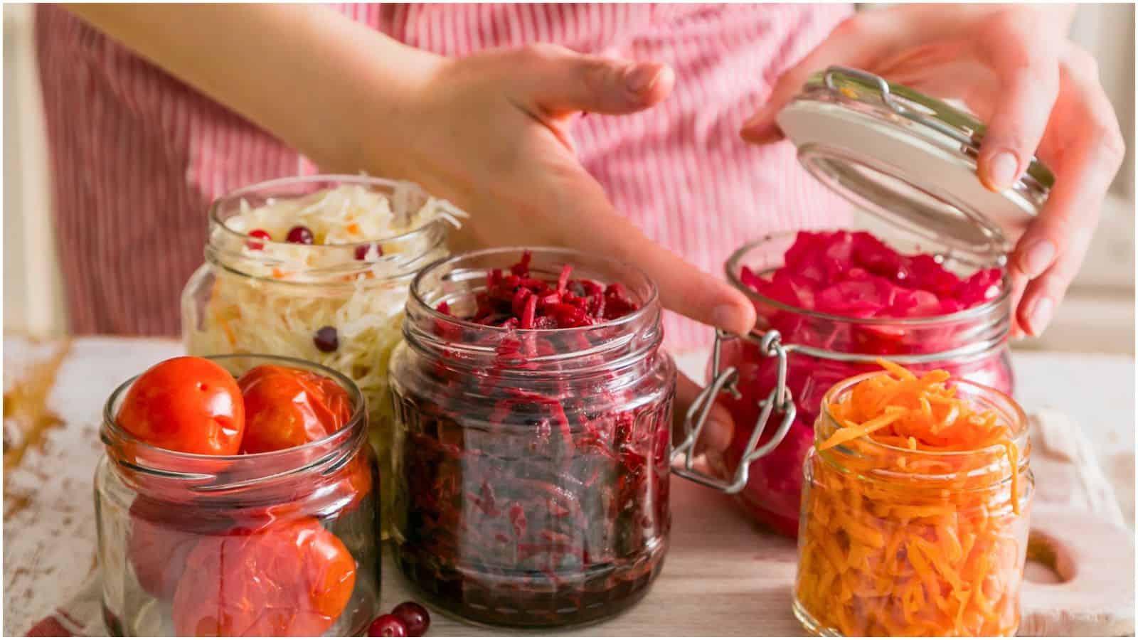 A person arranging and opening jars filled with various preserved vegetables, including shredded beets, cabbage, carrots, and whole tomatoes, on a wooden surface. Wearing a red-striped apron.