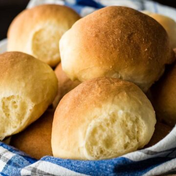 A basket filled with several round, golden-brown dinner rolls from treasured bread recipes. The rolls are wrapped in a blue and white checkered cloth, and the surface of the bread appears soft and fluffy. The background is dark, making the bread stand out.