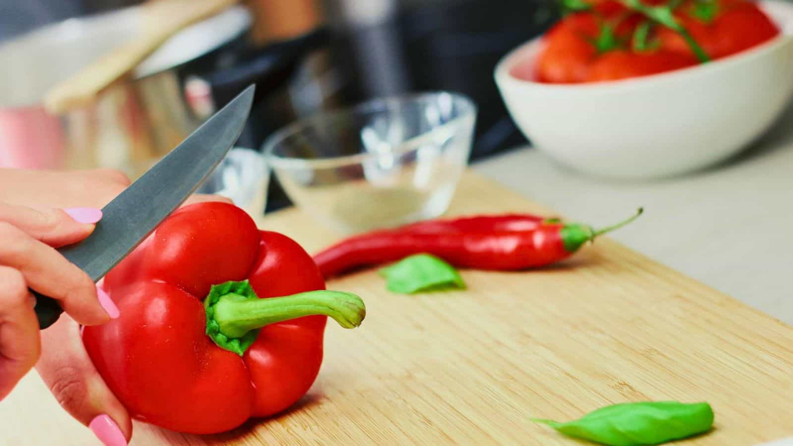A person is slicing a red bell pepper with a knife on a wooden cutting board. Other ingredients, including a red chili pepper and basil leaves, are also on the board.