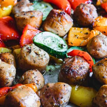 A close-up of a cooked dish featuring seasoned meatballs, sliced red and yellow bell peppers, and zucchini pieces. This easy summer dinner appears to be baked, with visible oil and herbs coating the ingredients.