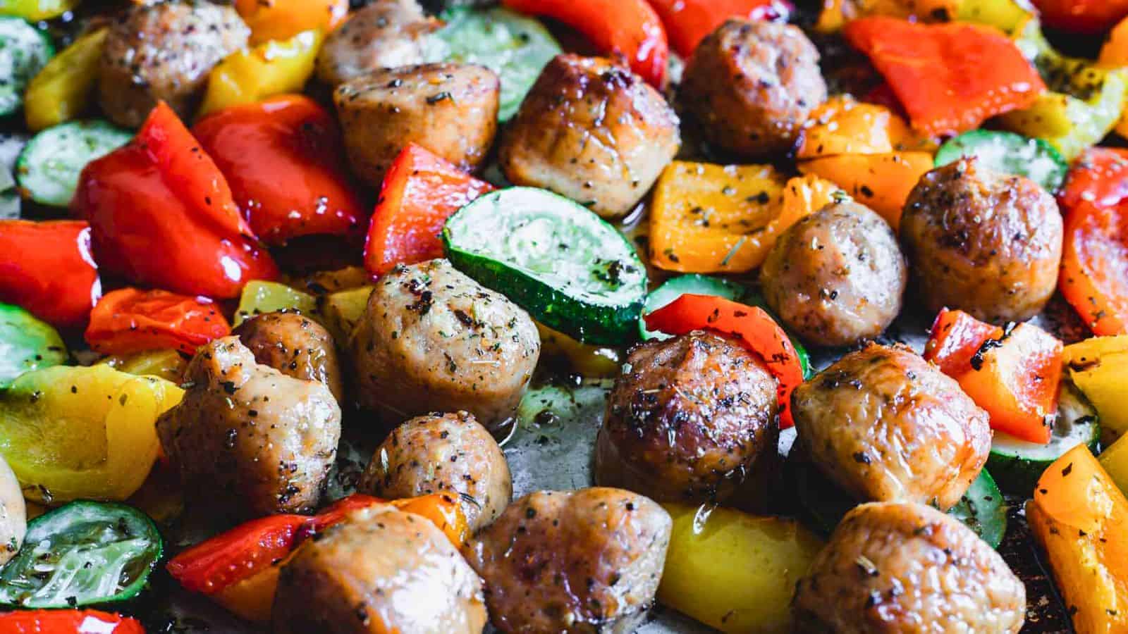 A close-up of a cooked dish featuring seasoned meatballs, sliced red and yellow bell peppers, and zucchini pieces. This easy summer dinner appears to be baked, with visible oil and herbs coating the ingredients.