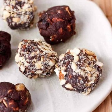 A white plate holds five chocolate truffles. Some are coated in chopped nuts, while others appear plain. The plate is set on a wooden surface, with mixed nuts, seeds, and dried berries scattered in the background.
